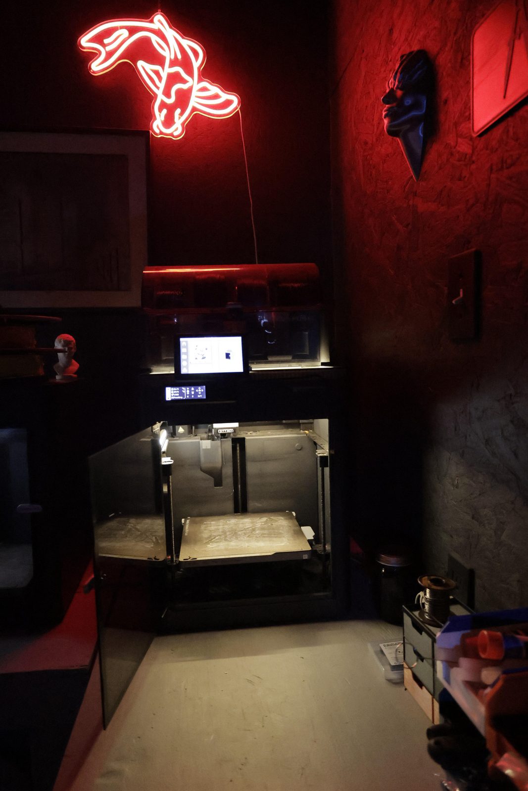 Close-up of an enclosed 3D printer on the TacticalTech3D bench, lit by the red koi neon sign on the wall above it.